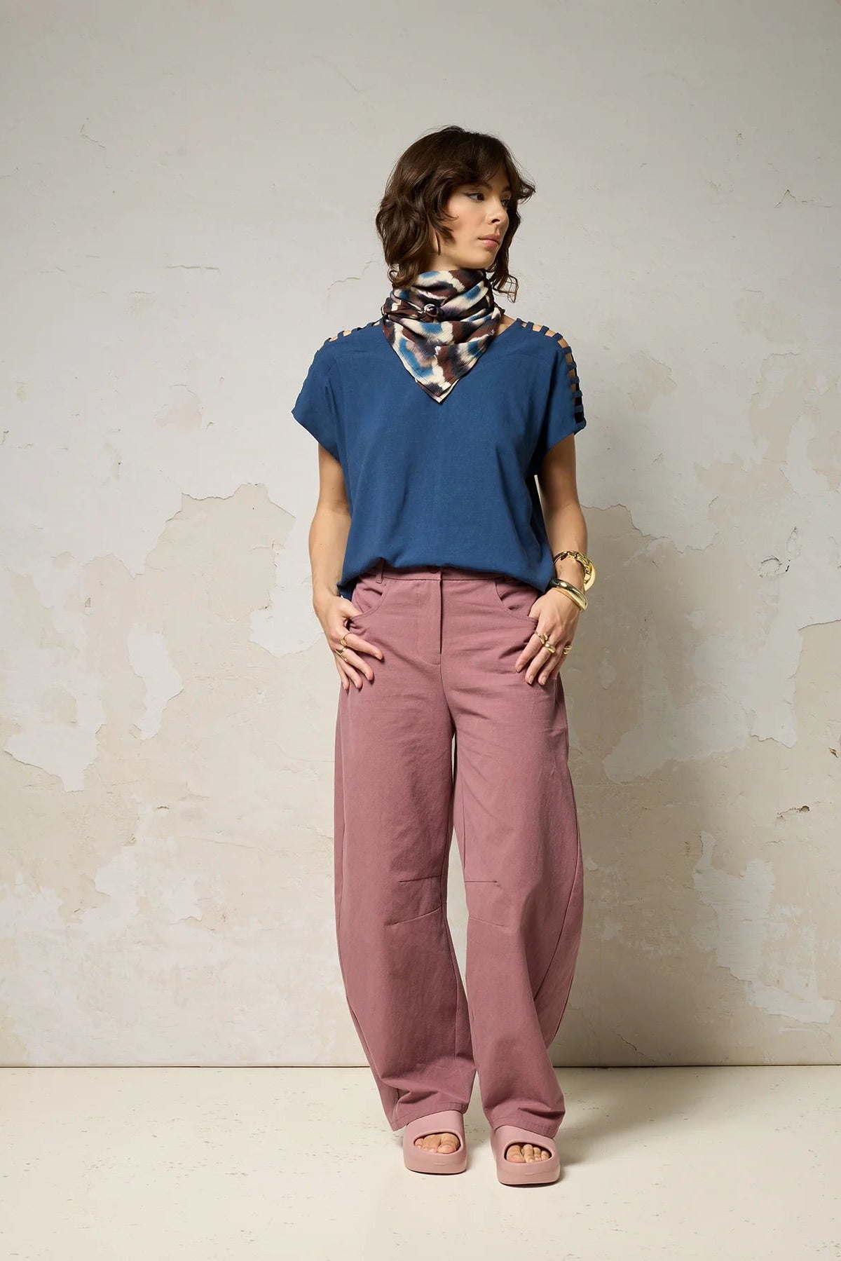 A woman wearing the Mia Top by Cokluch in Dahlia pink, a short sleeved top with openwork ladder-like cutouts at the shoulders, a loose fit, and V-neck. She is wearing it with black pants and standing against a plain background. 