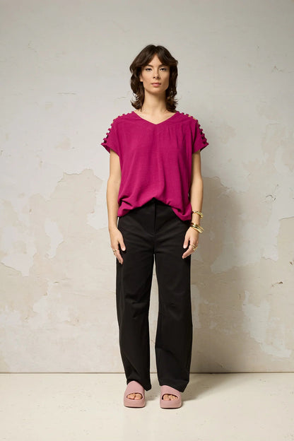 A woman wearing the Mia Top by Cokluch in Dahlia pink, a short sleeved top with openwork ladder-like cutouts at the shoulders, a loose fit, and V-neck. She is wearing it with black pants and standing against a plain background. 