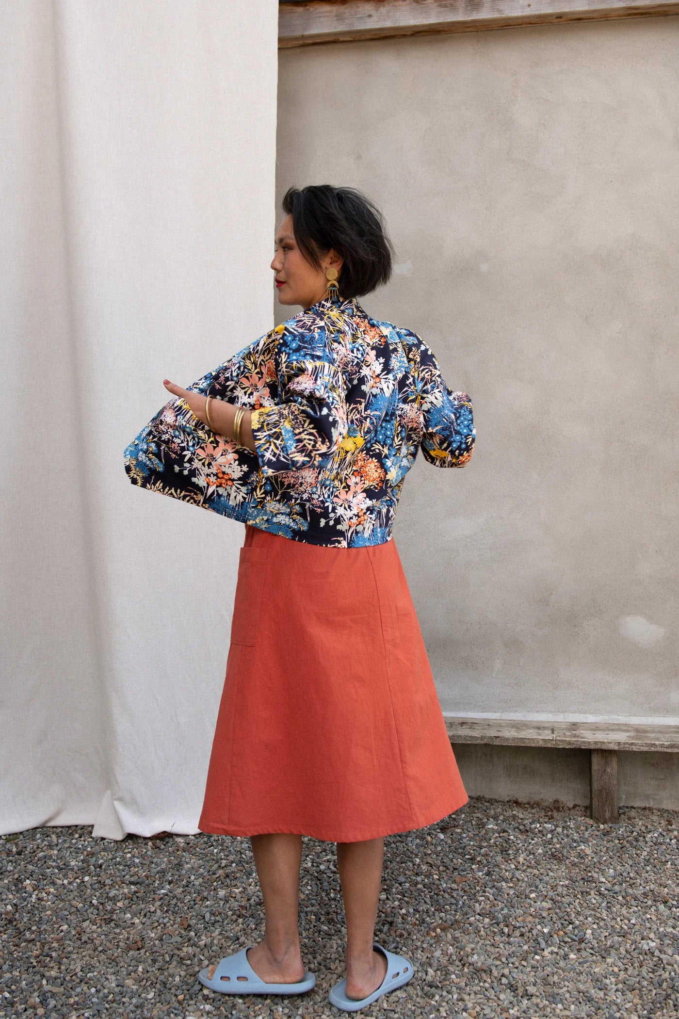 Back view of a woman wearing the Nymphea Screenprinted Skirt by Kazak in Red Pysanka with a floral jacket, she is standing outside in front of white curtain. 