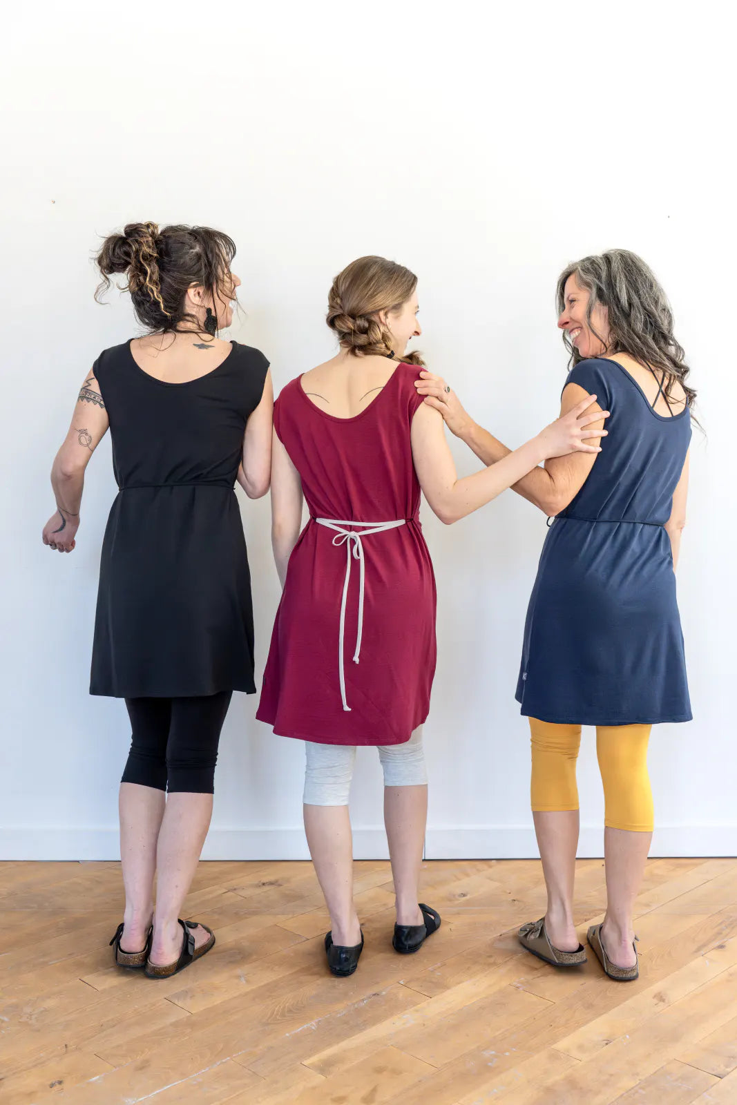 Back view of three women wearing the Mariposa Dress by Marie C, a short sleeved short dress that's reversible back to front with printed yoke on one side, and an attached tie belt. The one in the centre is wearing it in Raspberry. 