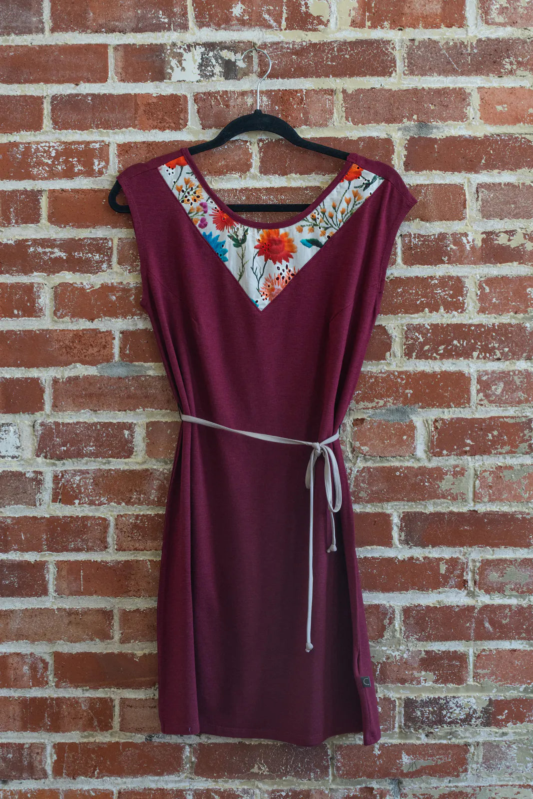 The Mariposa Dress by Marie C in Raspberry, a short sleeved short dress that's reversible back to front with printed yoke on one side, and an attached tie belt, is shown hanging on a brick wall. 