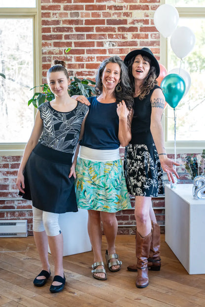 Three women wearing the Sol Top from Marie C, a reversible cap sleeve tee. From left to right they are wearing it in Black with Print, Navy, and Black. They are all wearing skirts and are standing at a party. 