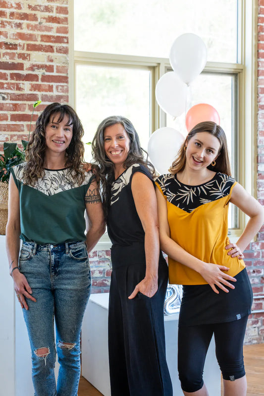 Three women wearing the Pepita Top from Marie C, a reversible t-shirt with a printed fabric yoke on one side. From the left they are wearing it in 
Green, Black and White, and Yellow, in a room with brick walls and balloons.