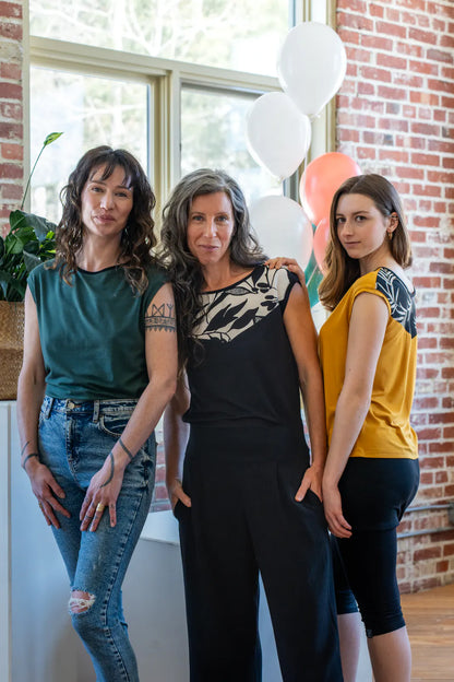 Three women wearing the Pepita Top from Marie C, a reversible t-shirt with a printed fabric yoke on one side. From the left they are wearing it in Green, Black and White, and Yellow. They are standing in a room with brick walls and balloons. 
