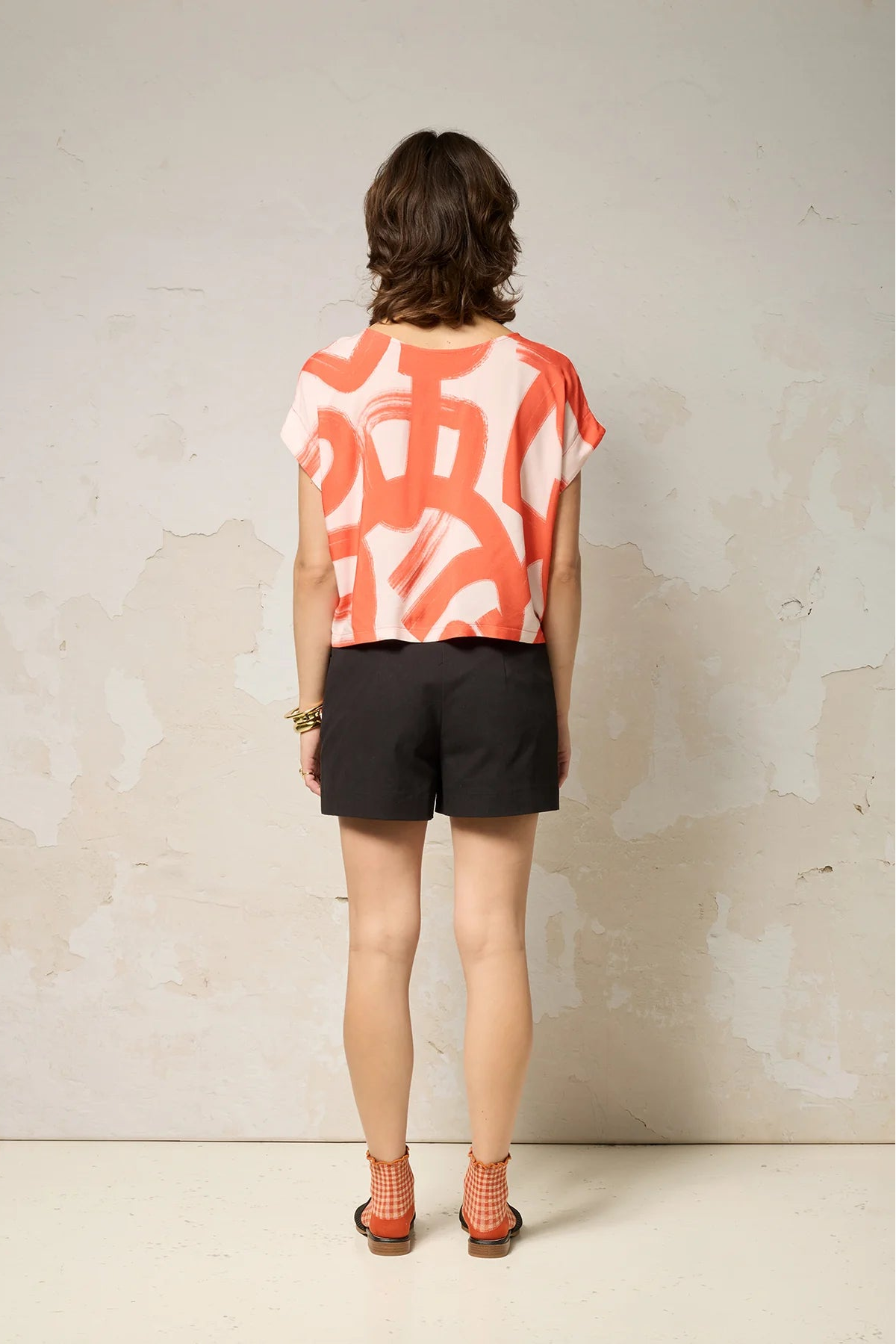 Back view of a woman wearing the Nyx top from Cockluch in Red Shodo, a short sleeved, loose fitting top with a drawstring detail at the bottom hem. She is wearing it with black shorts and standing in front of a textured background. 