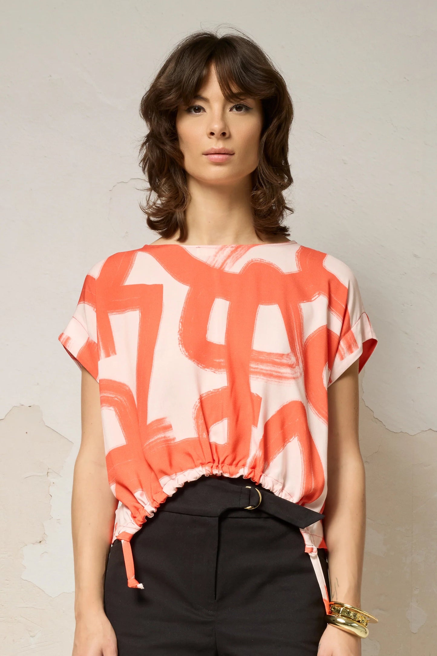 Waist up view of a woman wearing the Nyx top from Cockluch in Red Shodo, a short sleeved, loose fitting top with a drawstring detail at the bottom hem. She is wearing it with black shorts and standing in front of a textured background. 