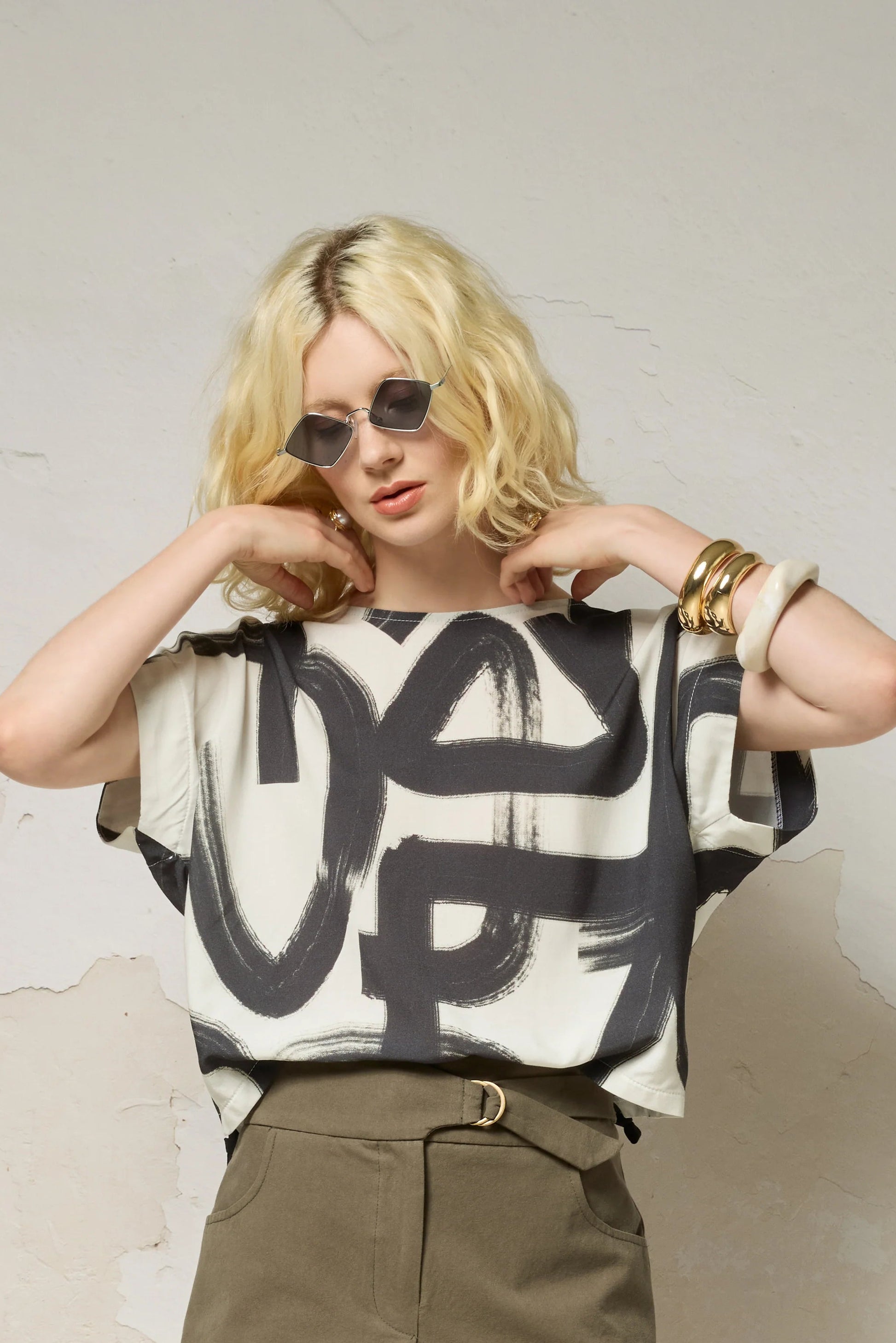 Waist up view of a woman wearing the Nyx top from Cockluch in Black Shodo, a short sleeved, loose fitting top with a drawstring detail at the bottom hem. She is wearing it with sunglasses and khaki shorts and standing in front of a textured background. 