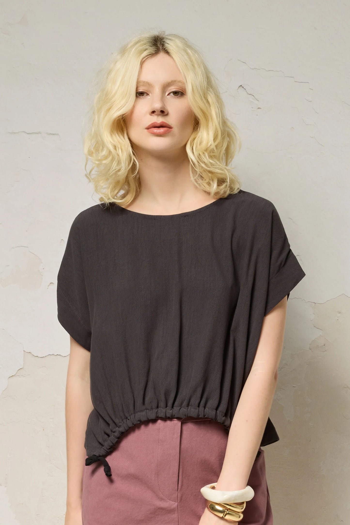 Waist up view of a woman wearing the Nyx top from Cockluch in Iron, a short sleeved, loose fitting top with a drawstring detail at the bottom hem. She is wearing it with pink shorts and standing in front of a textured background. 