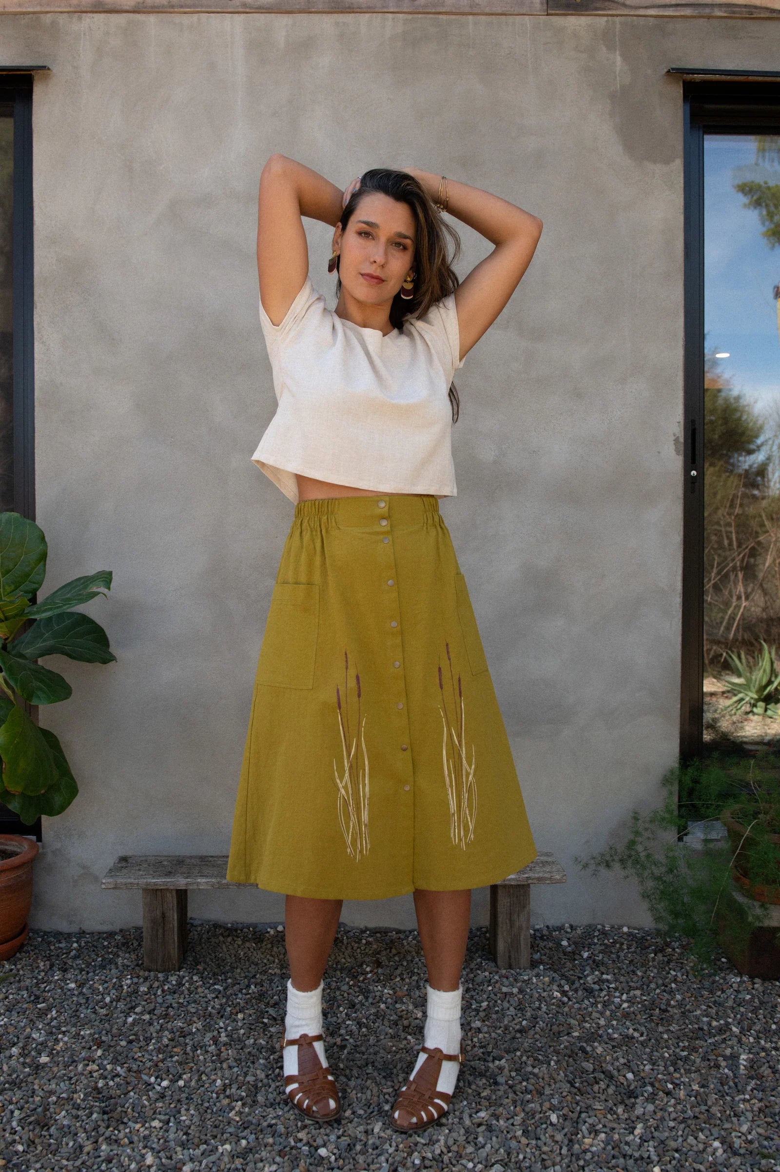 A woman wearing the Nymphea Screenprinted Skirt by Kazak in Green with Cattails, with a cream top. She is standing outside in front of a grey wall. 