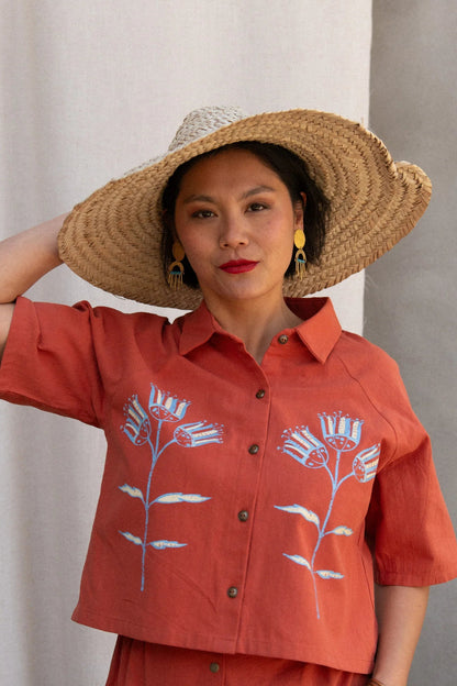 Waist up view of a woman wearing the Hosta Screen-printed Blouse by Kazak in Red with Pysanka, with hand-screen-printed flowers, mid-length raglan sleeves, a classic collar, and a button front. She is wearing it with a matching skirt and straw hat and standing outside by a grey wall. 