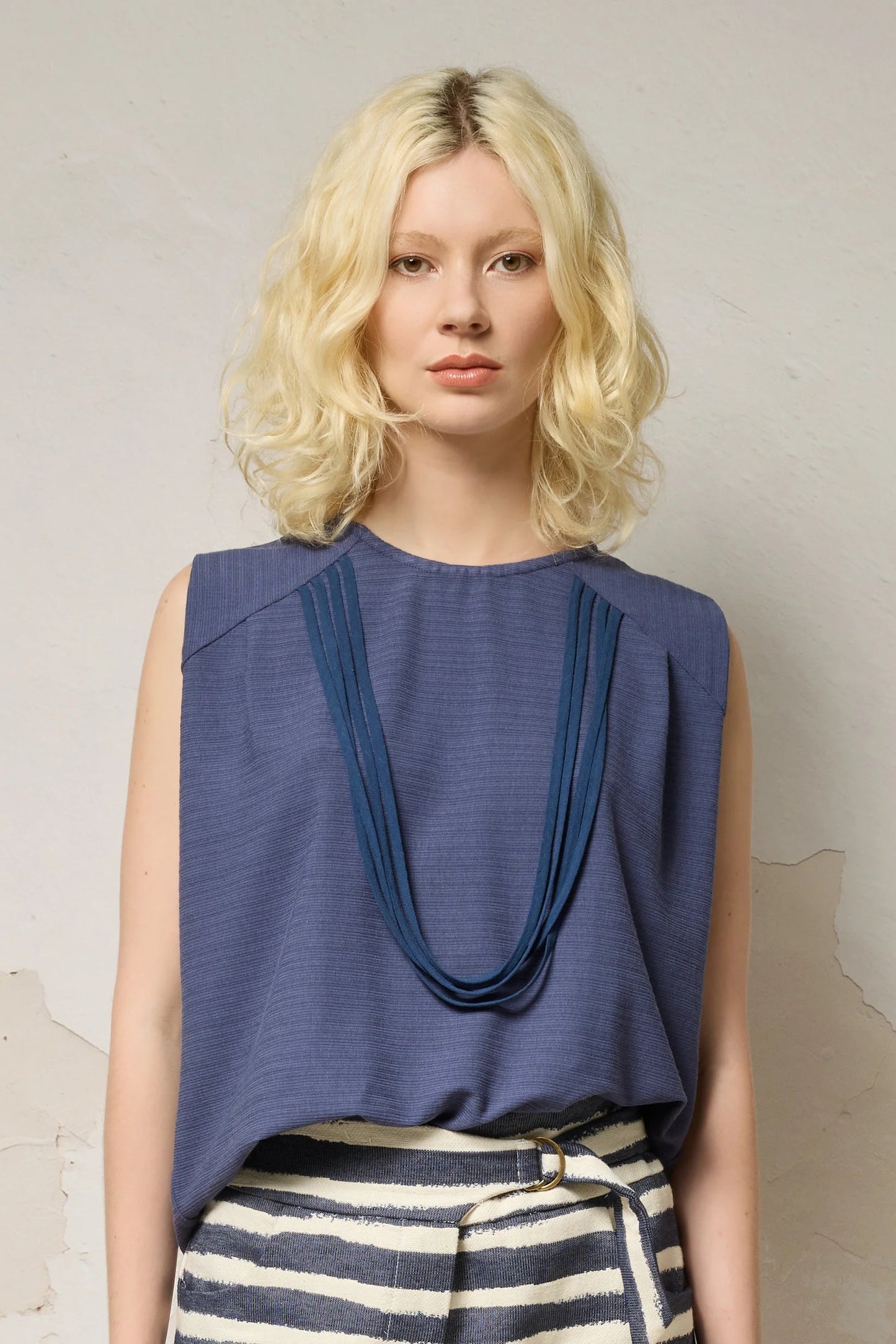 Waist up view of a woman wearing the Path Top from Cokluch in Black, a sleeveless top with line details at the shoulders and decorative straps that attach at each shoulder and hang to the front. She is wearing it with striped shorts and standing in front of a grey background. 
