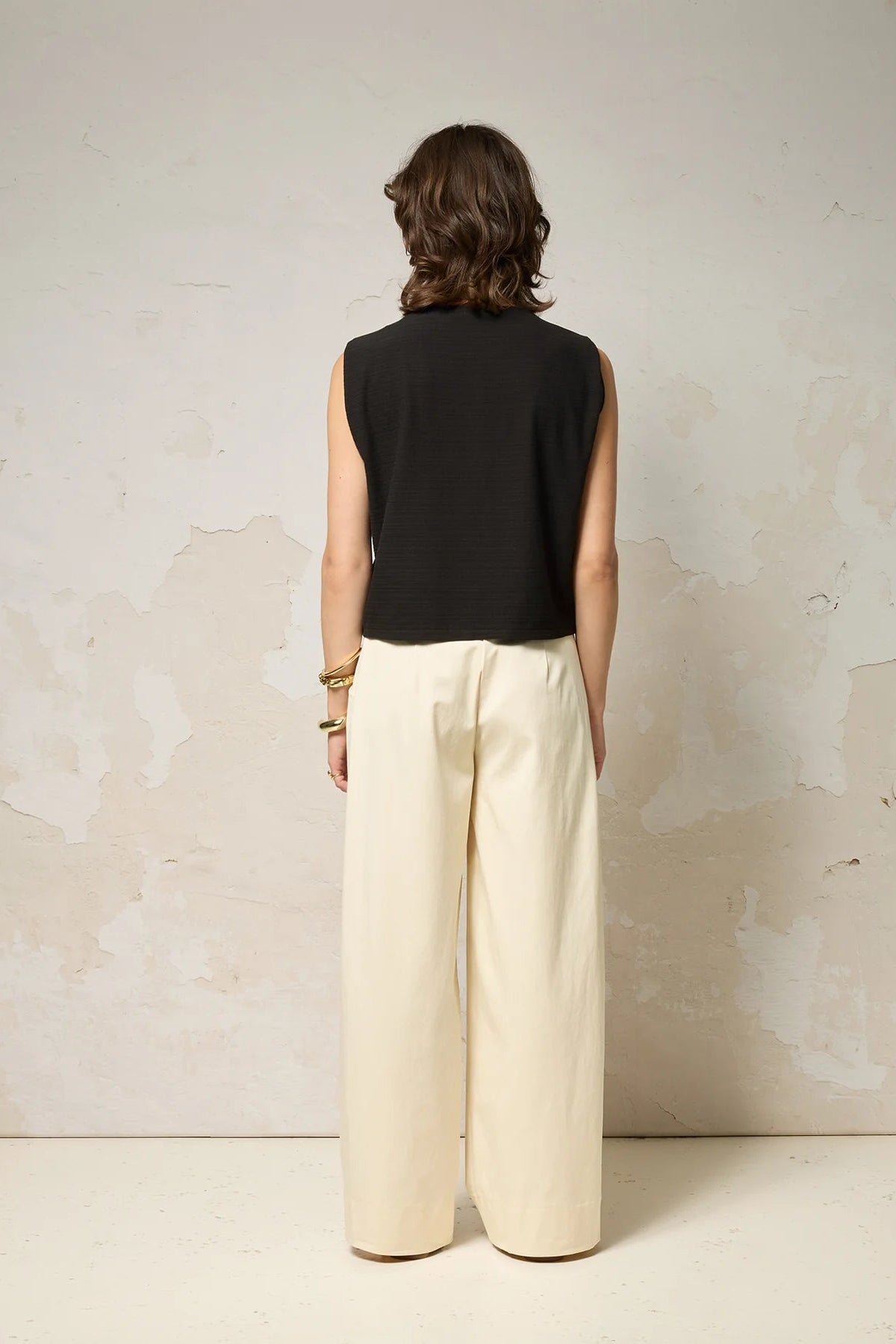 Back view of a woman wearing the Path Top from Cokluch in Black, a sleeveless top with line details at the shoulders and decorative straps that attach at each shoulder and hang to the front. She is wearing it with beige pants and a denim belt and standing in front of a grey background.