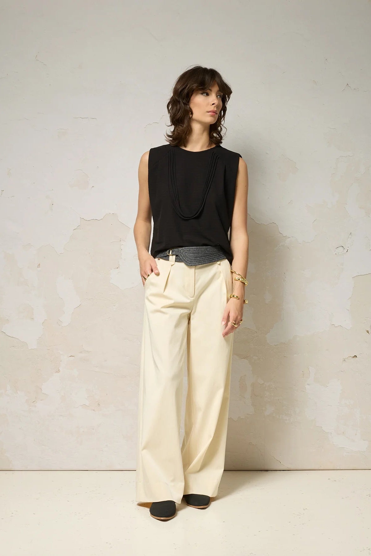 A woman wearing the Path Top from Cokluch in Black, a sleeveless top with line details at the shoulders and decorative straps that attach at each shoulder and hang to the front. She is wearing it with beige pants and a denim belt and standing in front of a grey background.