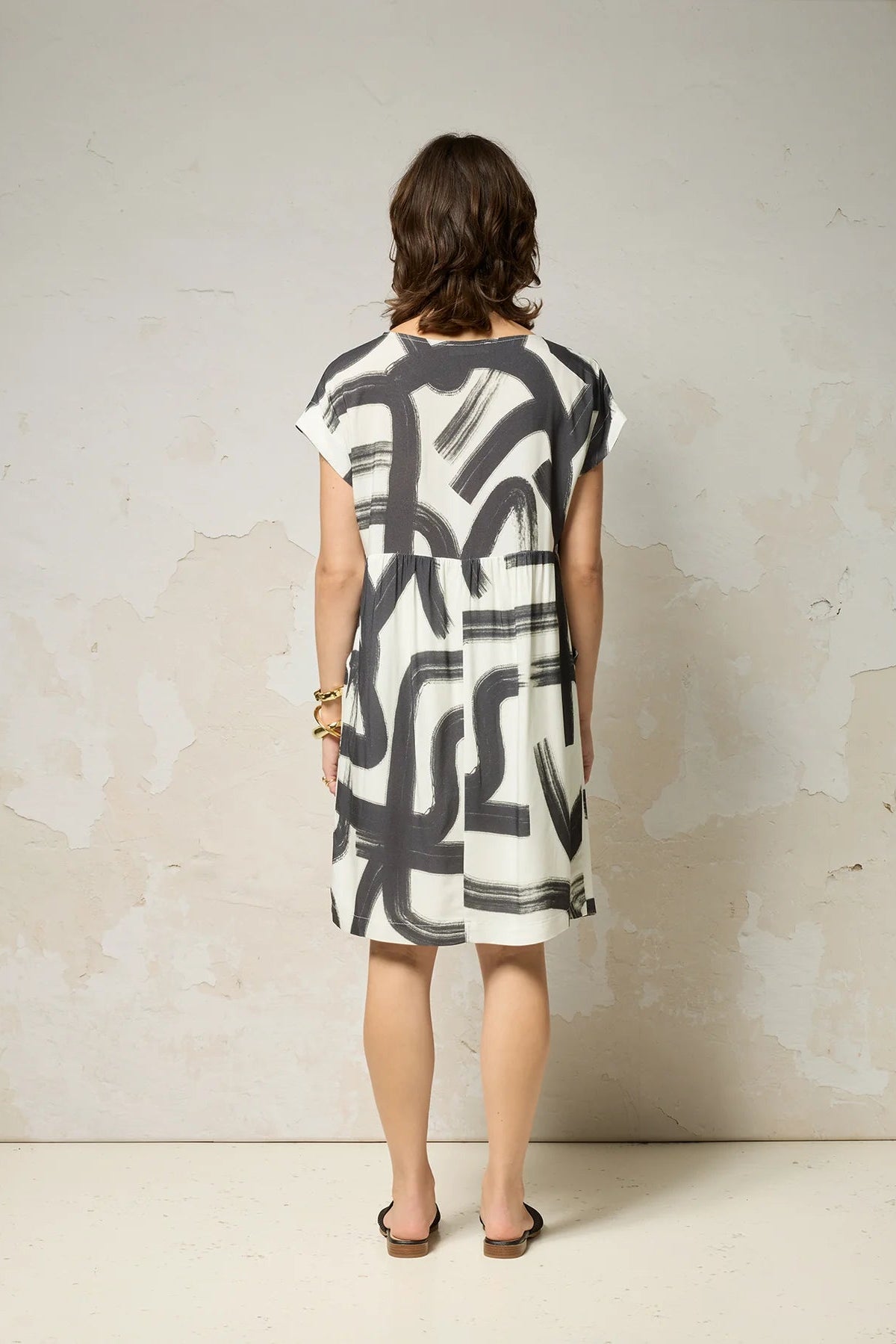 Back view of a woman wearing the Psyche Dress by Cokluch in Black Shodo print, a short sleeved loose fitting dress with a gathered waist, a triangle cut out detail at the front, and a knee-length hem. She is standing in front of a grey background. 