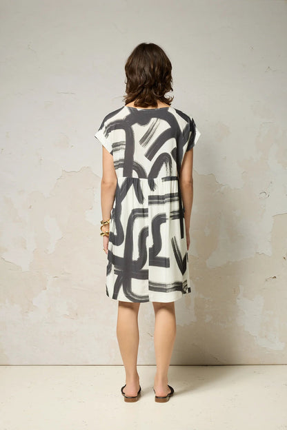 Back view of a woman wearing the Psyche Dress by Cokluch in Black Shodo print, a short sleeved loose fitting dress with a gathered waist, a triangle cut out detail at the front, and a knee-length hem. She is standing in front of a grey background. 