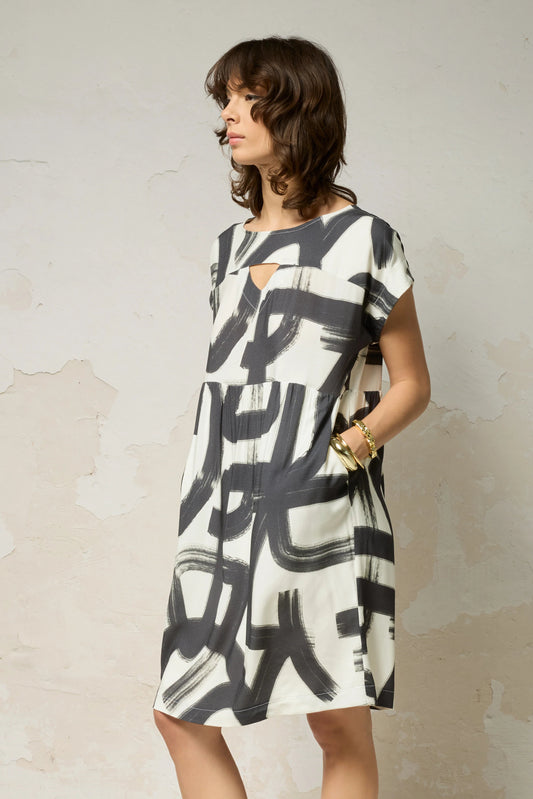 A woman wearing the Psyche Dress by Cokluch in Black Shodo print, a short sleeved loose fitting dress with a gathered waist, a triangle cut out detail at the front, and a knee-length hem. She is standing in front of a grey background. 