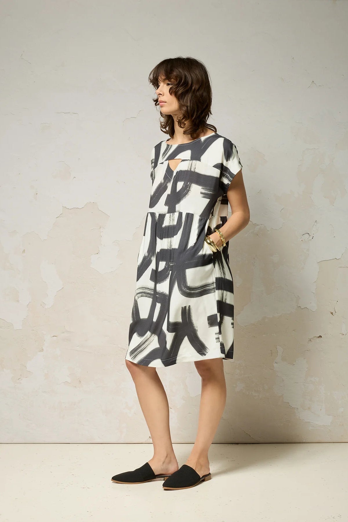 A woman wearing the Psyche Dress by Cokluch in Black Shodo print, a short sleeved loose fitting dress with a gathered waist, a triangle cut out detail at the front, and a knee-length hem. She is standing in front of a grey background. 