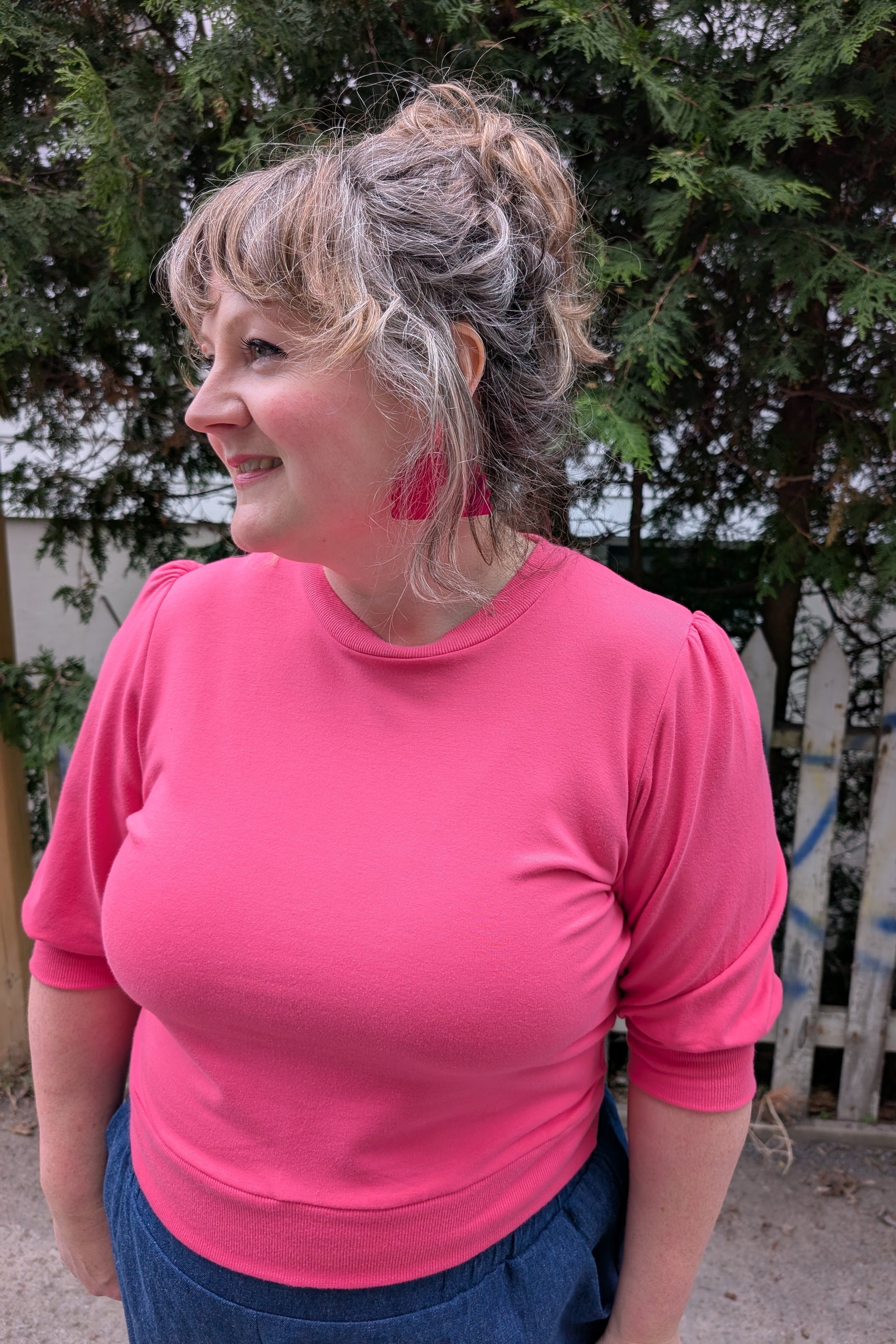 A woman wearing the Finch Sweatshirt by Allison Wonderland in Pink, a cropped sweatshirt with elbow-length puffed sleeves and ribbing at the neck and waistband. She is wearing it with denim pants and standing outside by a graffitied fence and trees.