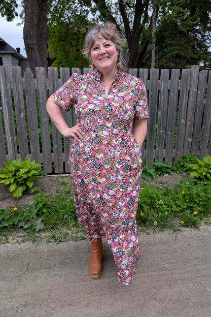 A woman wearing the Petal Print by Dotty in Pink/Blue floral,  a maxi dress with short flutter sleeves, a split V-neck and ruffled collar, with a gathered waist and A-line skirt. She is standing outside by a garden and a wooden fence. 