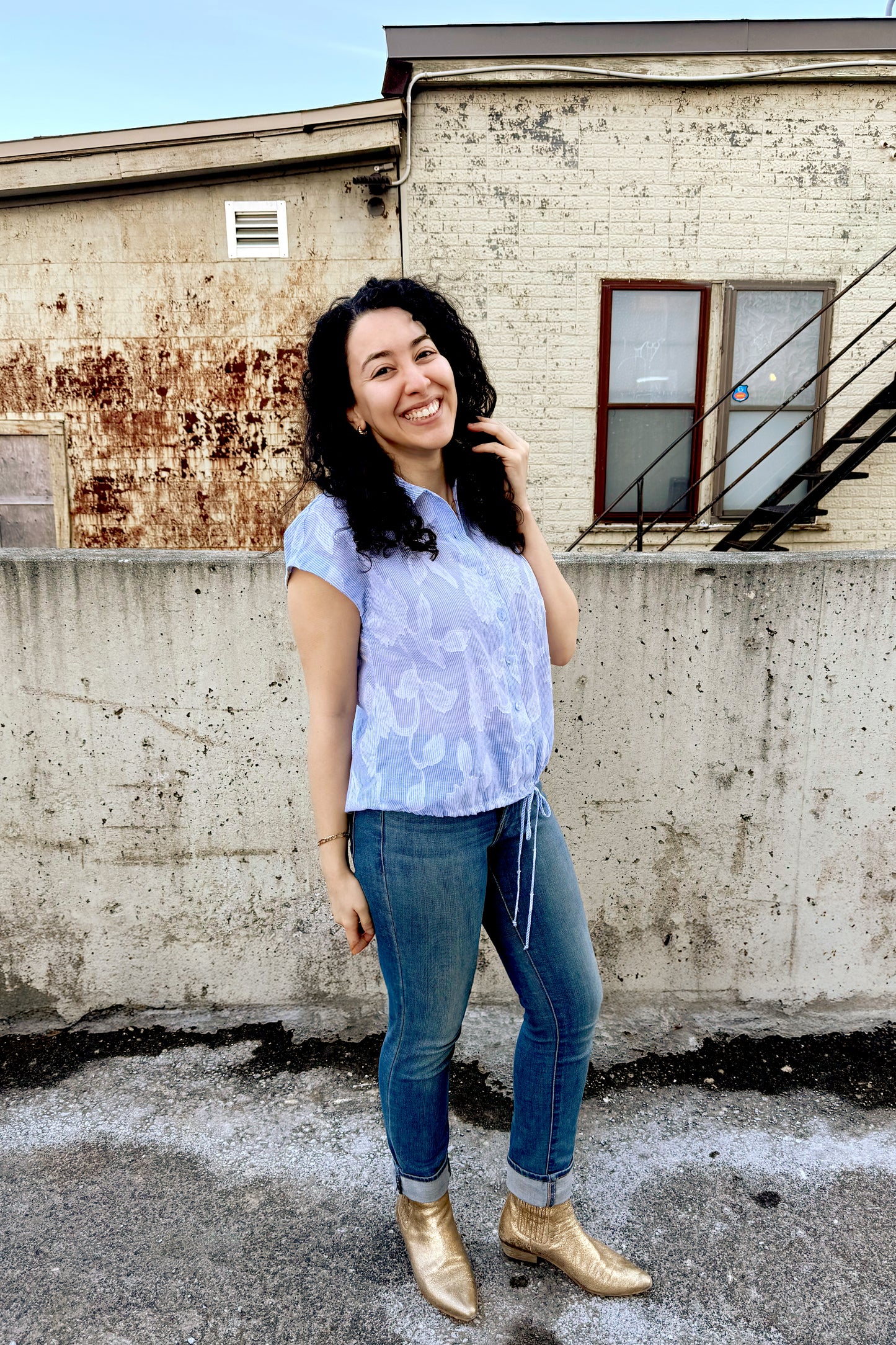 A woman wearing the Griffin Top by Pure Essence in Blue/White print, with a textured stripe/lace print, a short-sleeved blouse with a classic collar, button front, and drawstring detail at the waist.She is wearing it with jeans and standing outside by a brick wall. 