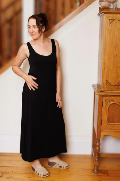 A woman wearing the Ruby Dress by Moovment in Black, a long camisole dress with side slits. She is standing against a white wall beside a wooden cupboard. 