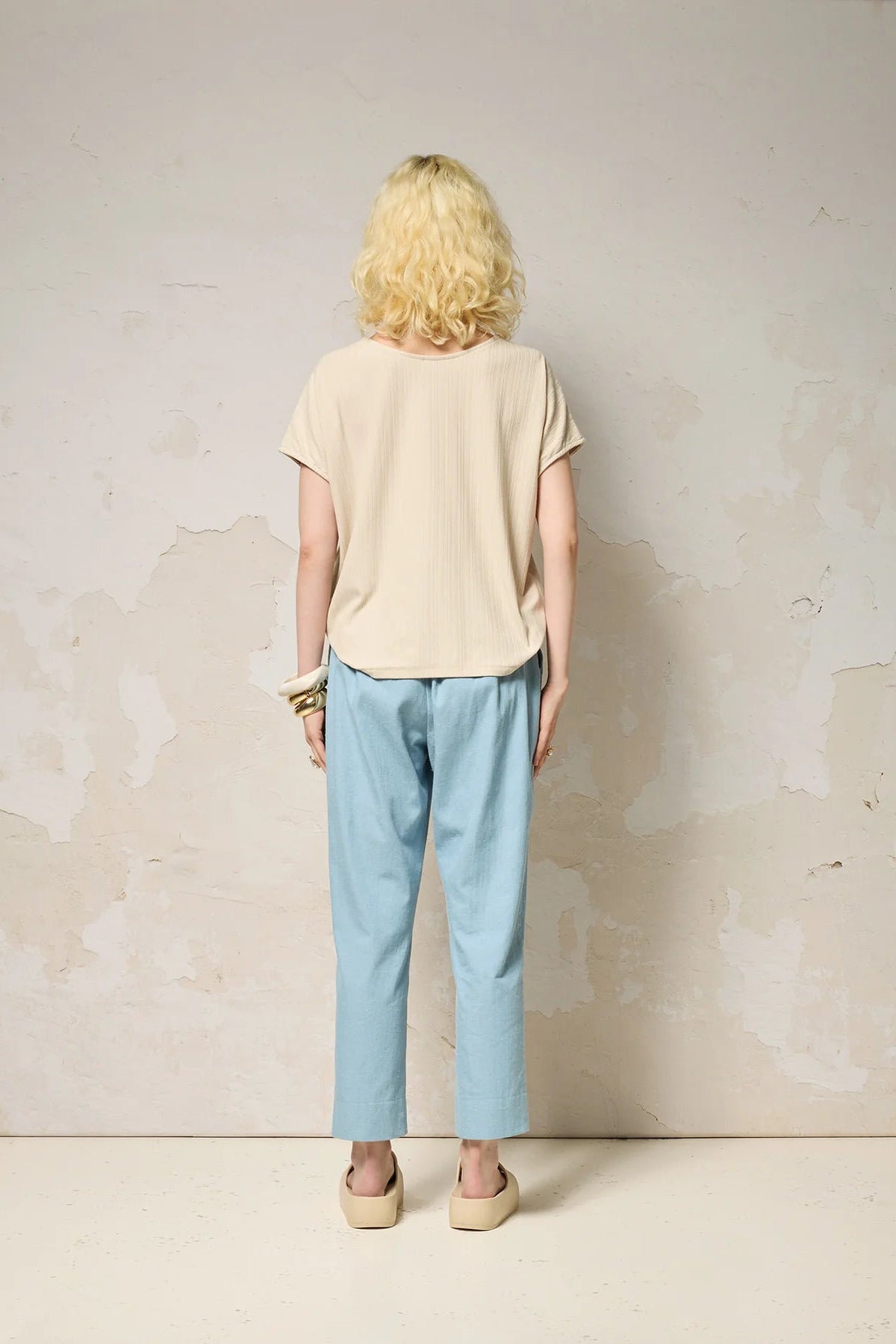 Back view of a woman wearing the Samsara Top by Cokluch in Sand, a short sleeved top with fringed V-panel at the chest and a relaxed fit. She is wearing it with blue pants and standing against a neutral background