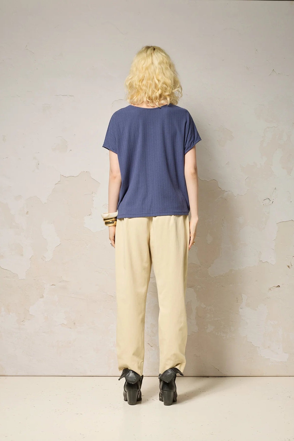 Back view of a woman wearing the Samsara Top by Cokluch in Navy, a short sleeved top with fringed V-panel at the chest and a relaxed fit. She is wearing it with cream coloured pants and standing against a neutral background