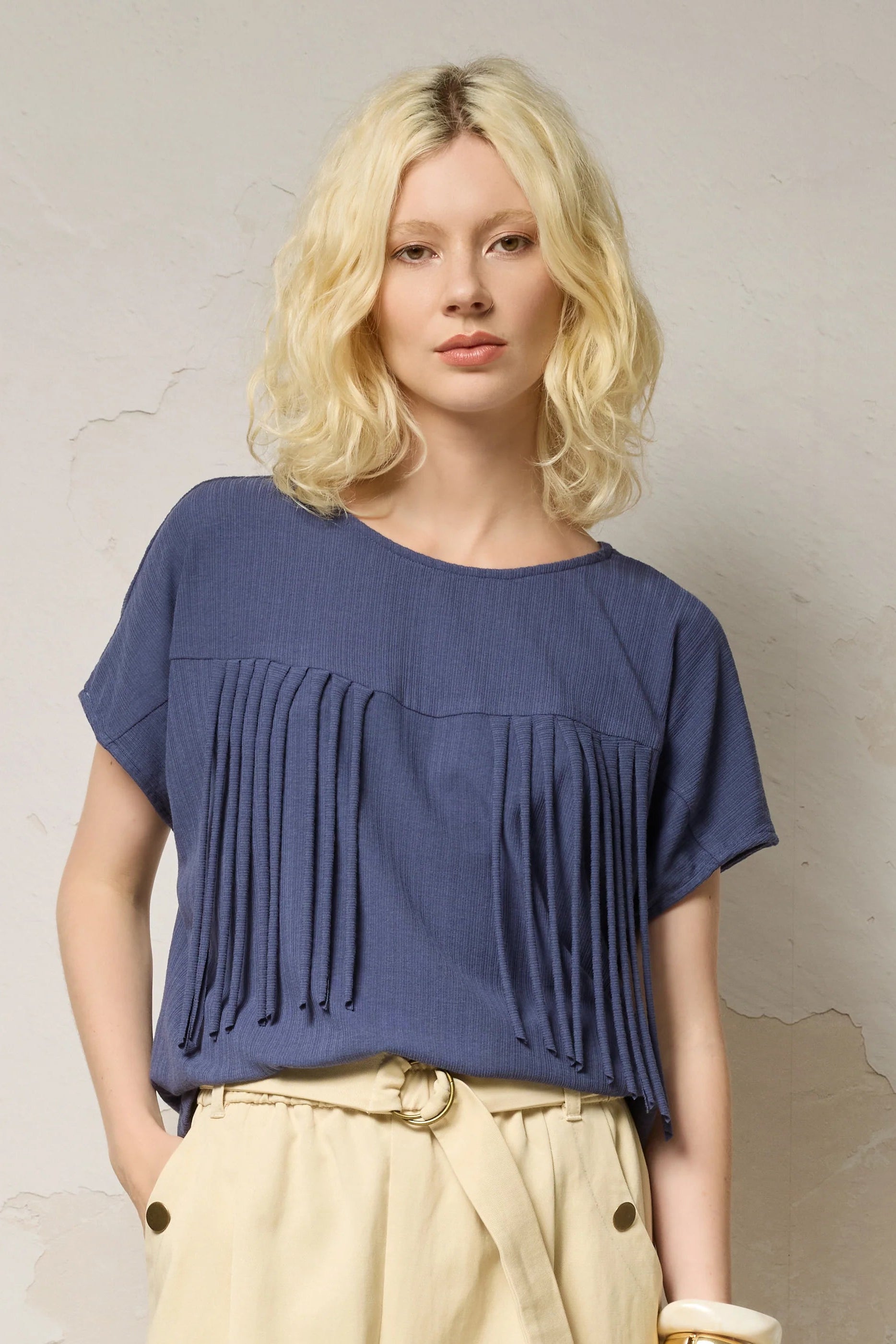 Waist up view of a woman wearing the Samsara Top by Cokluch in Navy, a short sleeved top with fringed V-panel at the chest and a relaxed fit. She is wearing it with cream coloured pants and standing against a neutral background