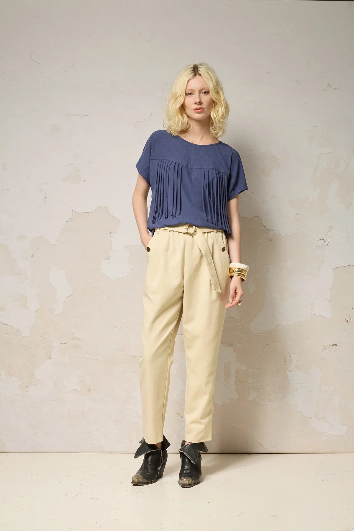 A woman wearing the Samsara Top by Cokluch in Navy, a short sleeved top with fringed V-panel at the chest and a relaxed fit. She is wearing it with cream coloured pants and standing against a neutral background