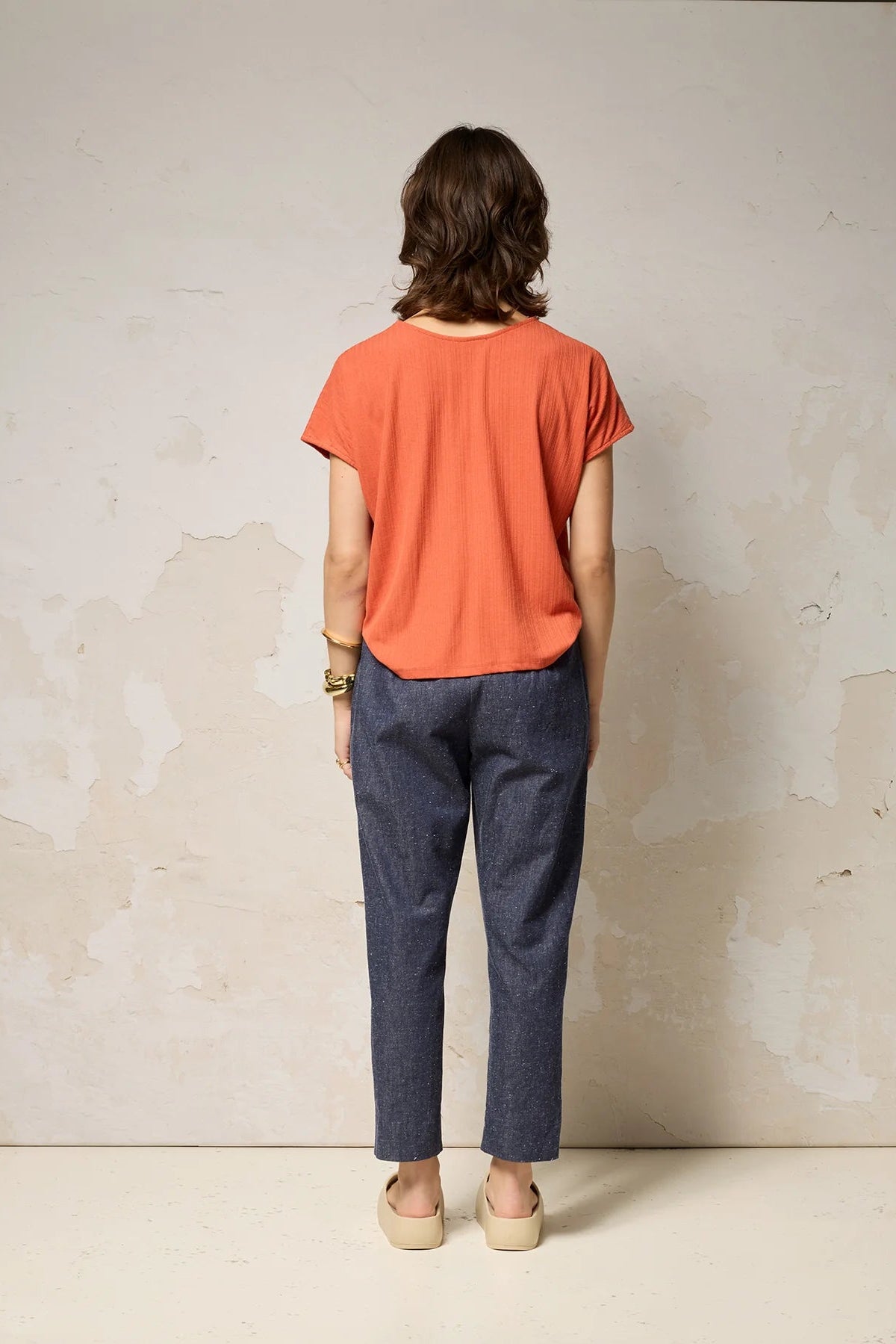 Back view of a woman wearing the Shiva Pants from Cokluch in Indigo Speckle, barrel legged pants in lightweight textured denim with an elastic waist, metal ring belt, and decorative buttons on the pockets. She is wearing them with a coral top and standing in front of a grey background. 