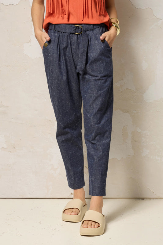 Waist down view of a woman wearing the Shiva Pants from Cokluch in Indigo Speckle, barrel legged pants in lightweight textured denim with an elastic waist, metal ring belt, and decorative buttons on the pockets. She is wearing them with a coral top and standing in front of a grey background. 