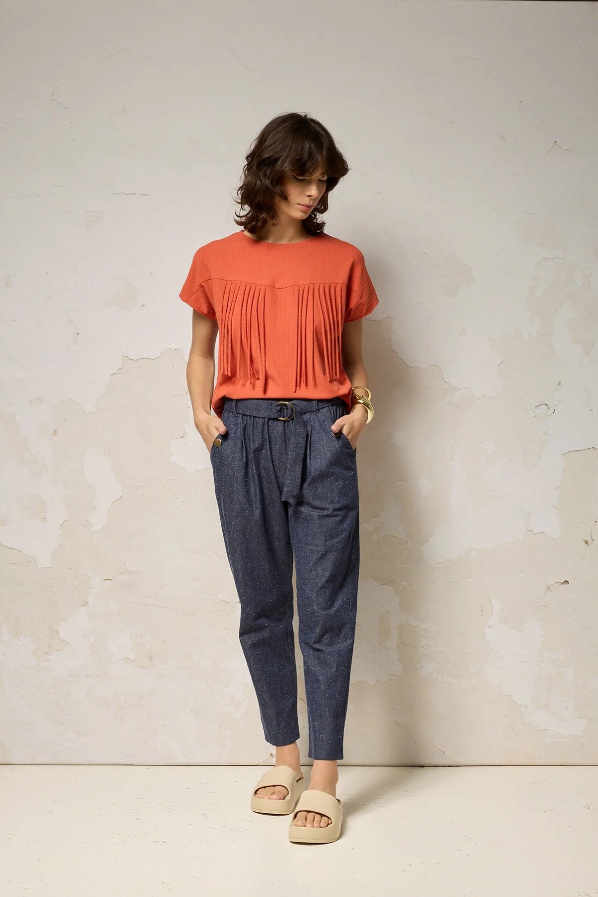 A woman wearing the Shiva Pants from Cokluch in Indigo Speckle, barrel legged pants in lightweight textured denim with an elastic waist, metal ring belt, and decorative buttons on the pockets. She is wearing them with a coral top and standing in front of a grey background. 