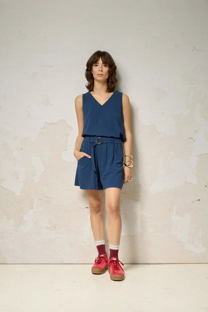 A woman wearing the Atman Shorts by Cokluch in Denim blue, mid-thigh length shorts with a high elastic waist and an attached ring belt. She is wearing them with a matching top and standing in front of a plain background. 