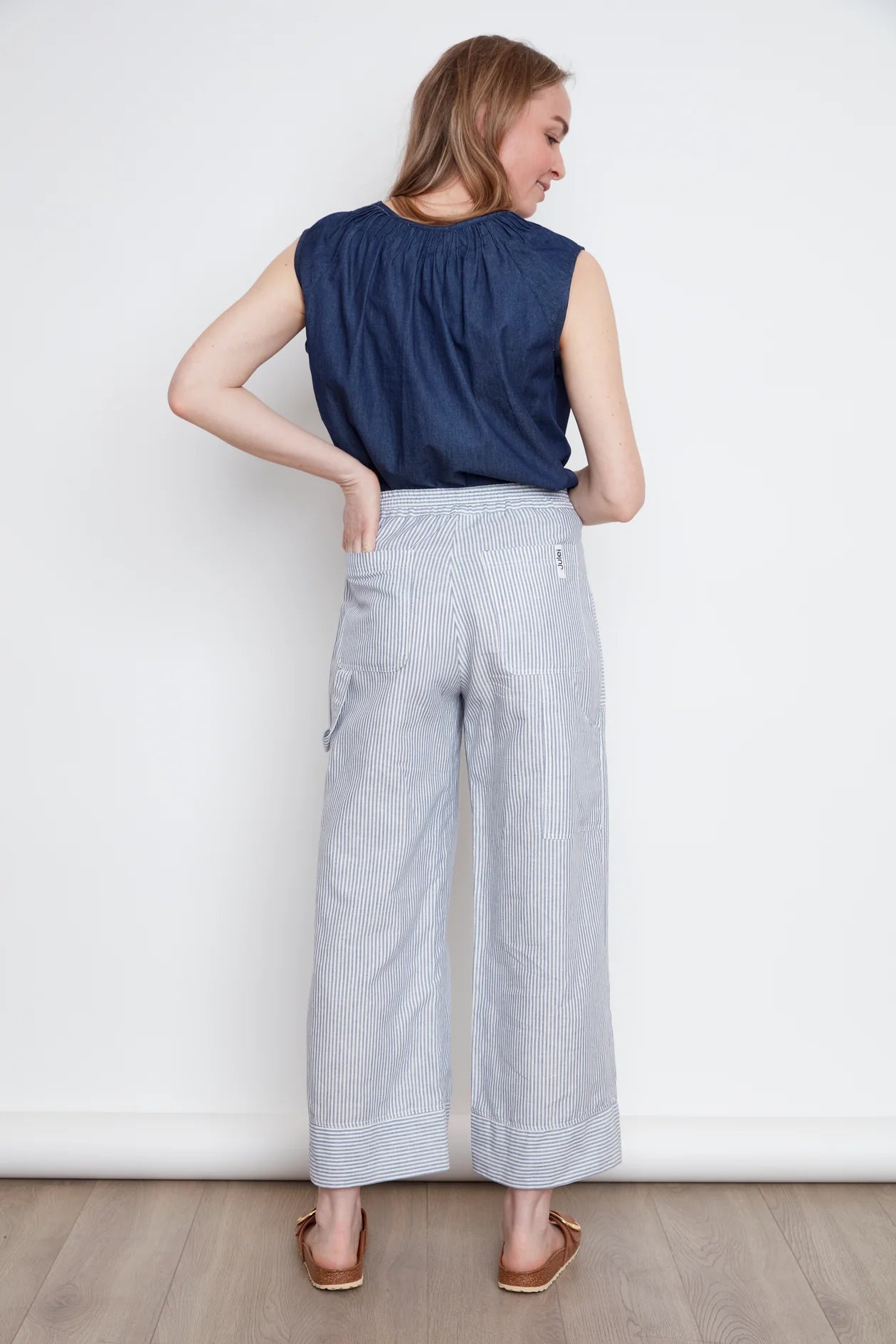 Back view of a woman wearing the Sabrina Top by Julei in Denim, with raglan cap sleeves, a notched v-neck with pleat details, and a pleated back yoke. She is wearing it with striped pants and standing against a white background