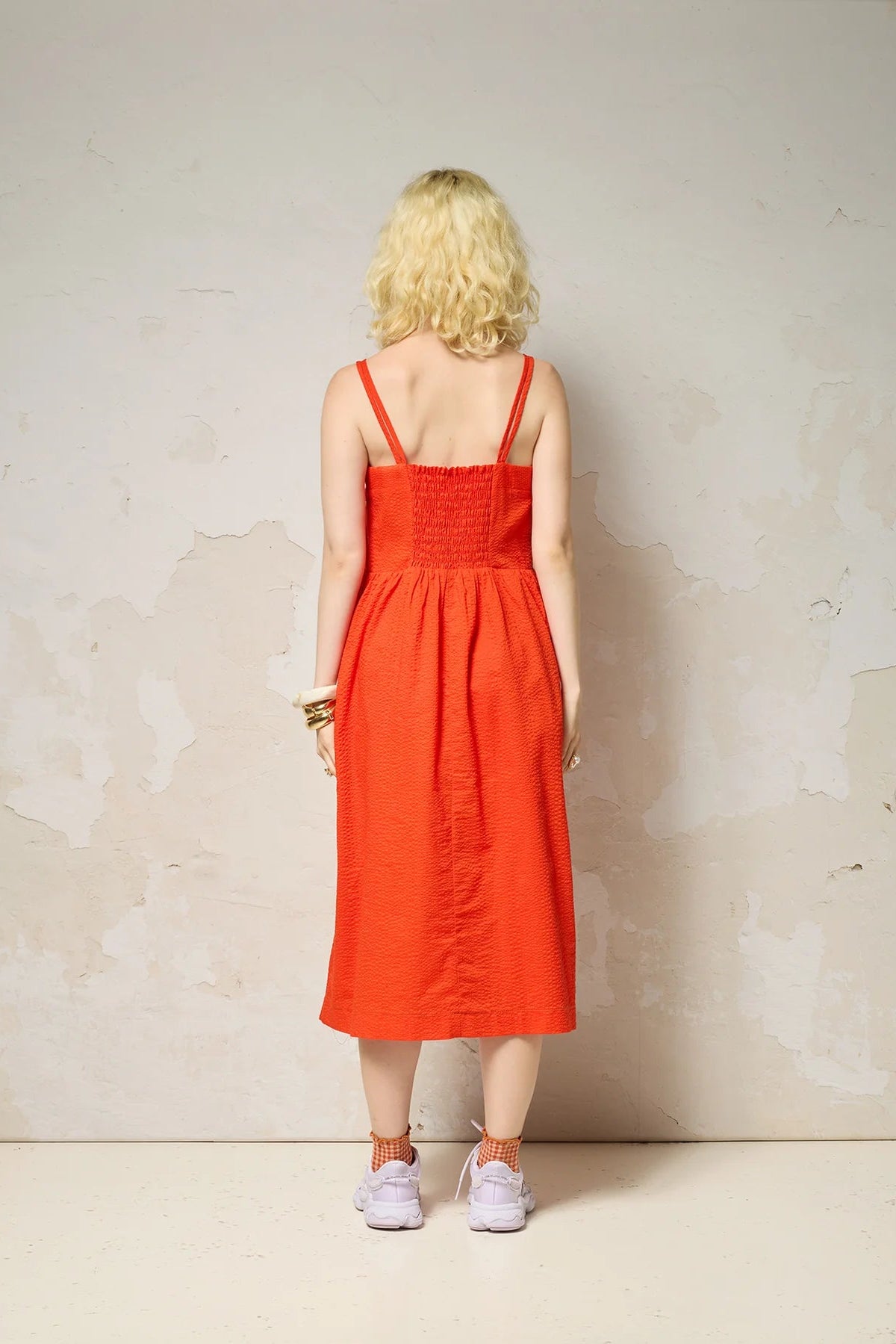 Back view of a woman wearing the Thalassa Dress by Cokluch in Fiesta, a midi dress with double spaghetti straps, a sweetheart neckline, and elastic smocking at the back, all in waffle textured cotton. She is standing against a plain background