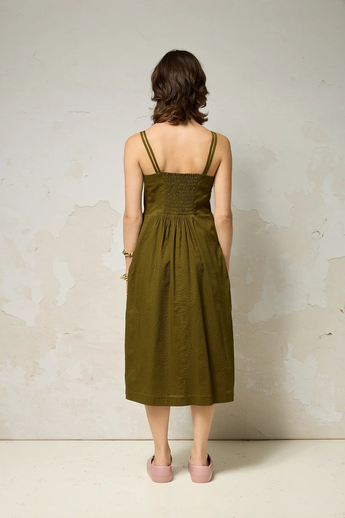 Back view of a woman wearing the Thalassa Dress by Cokluch in Olive, a midi dress with double spaghetti straps, a sweetheart neckline, and elastic smocking at the back, all in waffle textured cotton. She is standing against a plain background