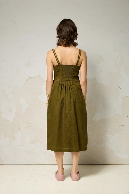 Back view of a woman wearing the Thalassa Dress by Cokluch in Olive, a midi dress with double spaghetti straps, a sweetheart neckline, and elastic smocking at the back, all in waffle textured cotton. She is standing against a plain background