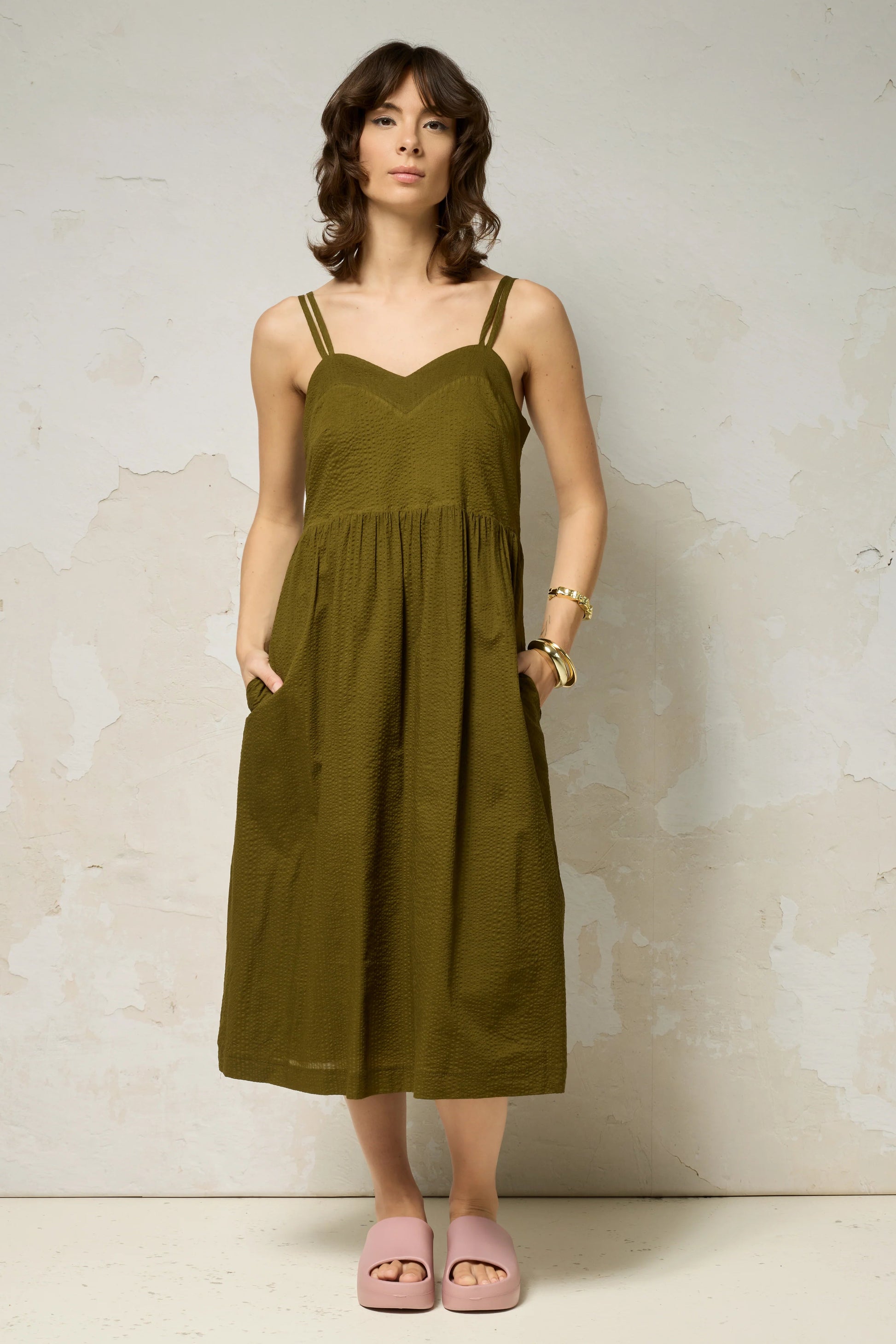 A woman wearing the Thalassa Dress by Cokluch in Olive, a midi dress with double spaghetti straps, a sweetheart neckline, and elastic smocking at the back, all in waffle textured cotton. She is standing against a plain background