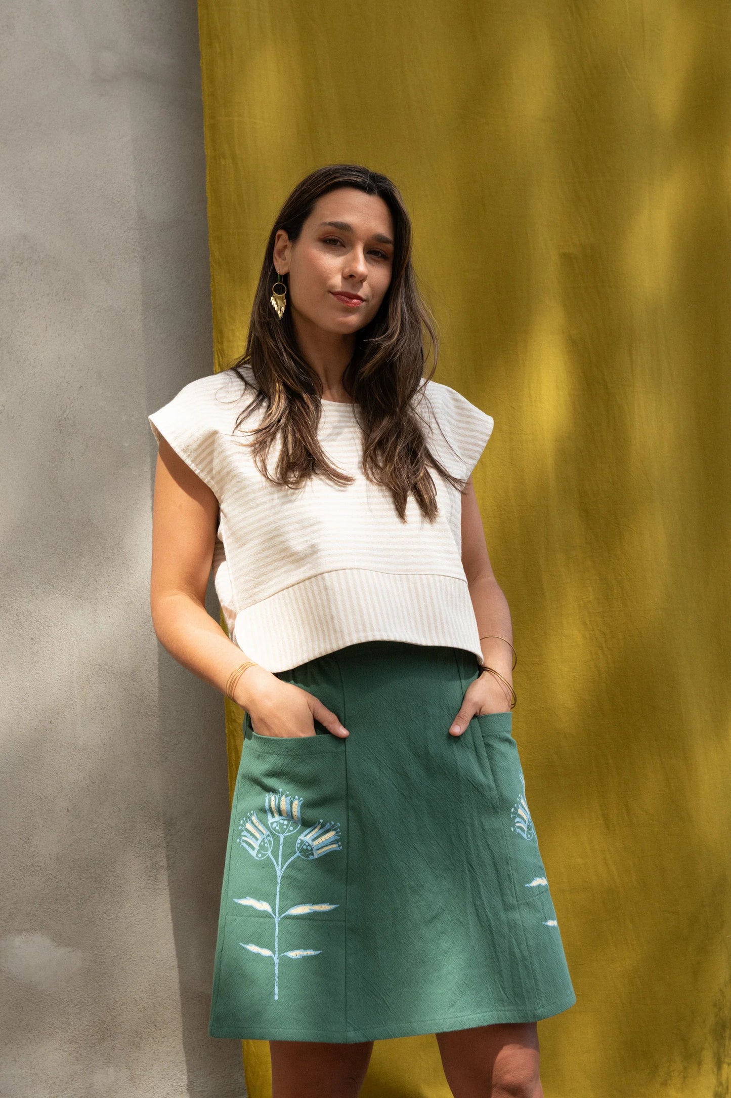 A woman wearing the Vicenze Screen-printed Skirt by Kazak in Green with Pysanka, an above the knee A-line skirt with hand-screened flowers, an elasticated waist, and large patch pockets. She is wearing it with the Tulip top in Taupe and standing outside by a grey wall and yellow curtains and trees.
