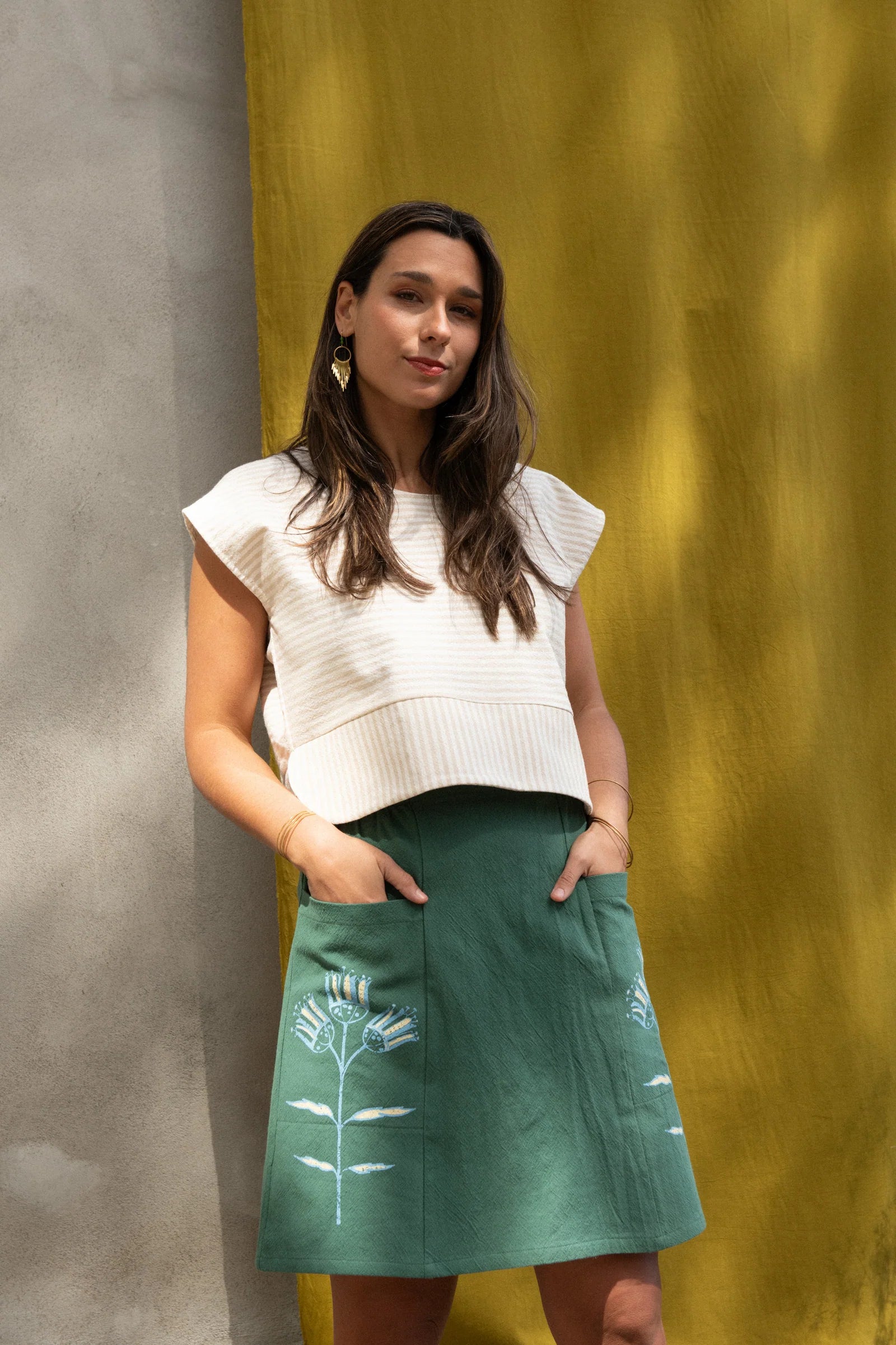 A woman wearing the Vicenze Screen-printed Skirt by Kazak in Green with Pysanka, an above the knee A-line skirt with hand-screened flowers, an elasticated waist, and large patch pockets. She is wearing it with the Tulip top in Taupe and standing outside by a grey wall and yellow curtains and trees.