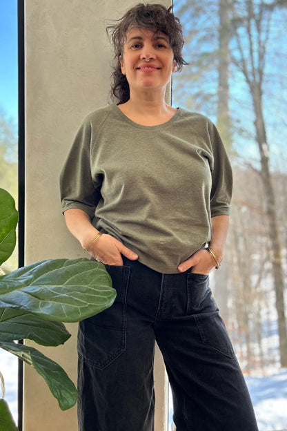 A woman wearing the Vernus Top by Kazak in Sage with elbow-length raglan sleeves with a pleat detail at the shoulders, a round neck, and a fit that tapers at the waist. She is wearing it with dark jeans standing indoors by a window.