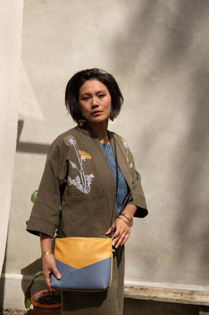 Side view of a woman wearing the Magnolia Screen-printed Jacket from Kazak in Olive with Dandelions, a short kimono style jacket with hand-screen printed dandelions, open front, 3/4-length sleeves, and big patch pockets. She is wearing it with matching pants and a blue and yellow purse and standing outside by a grey wall.