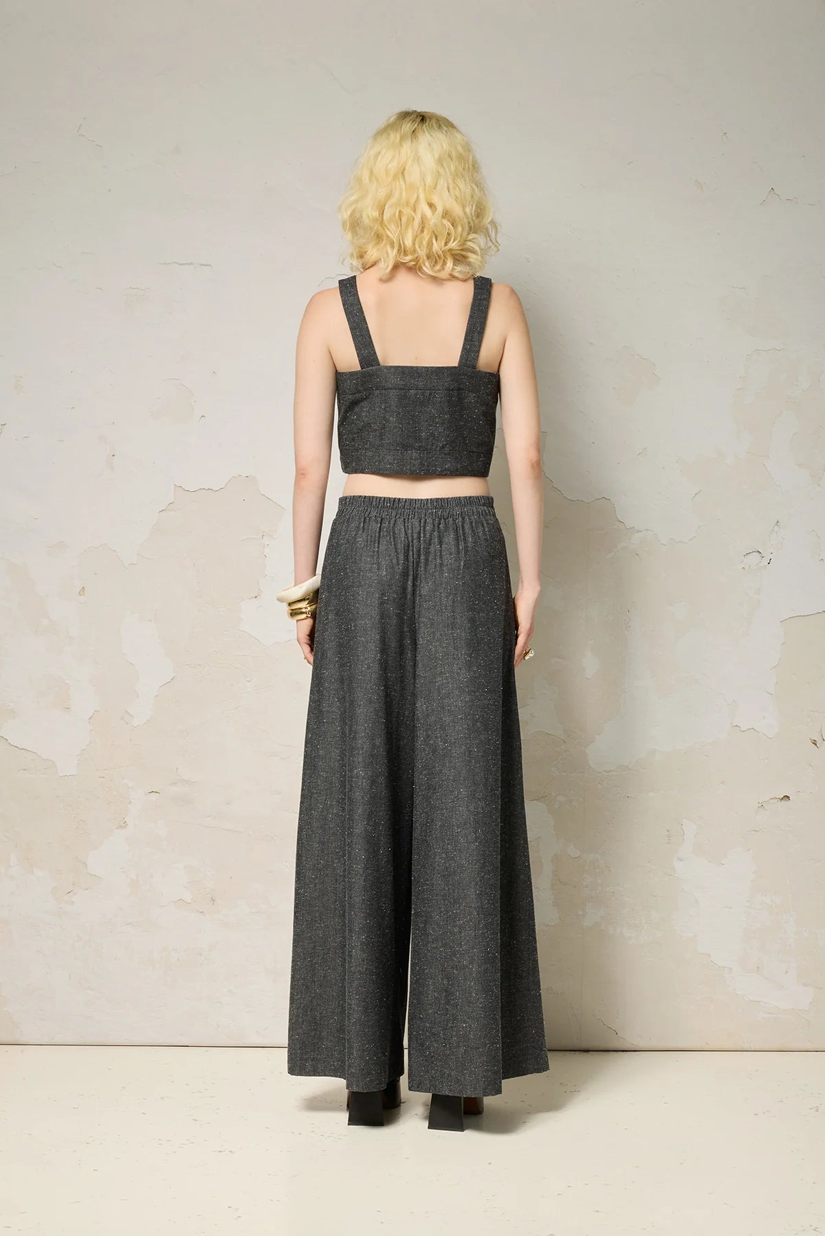 Back view of a woman wearing the Rama Pants by Cokluch in Black Speckle, textured denim pants with wide legs and a smooth elastic waistband. She is wearing them with a matching top and standing in front of a grey wall. 