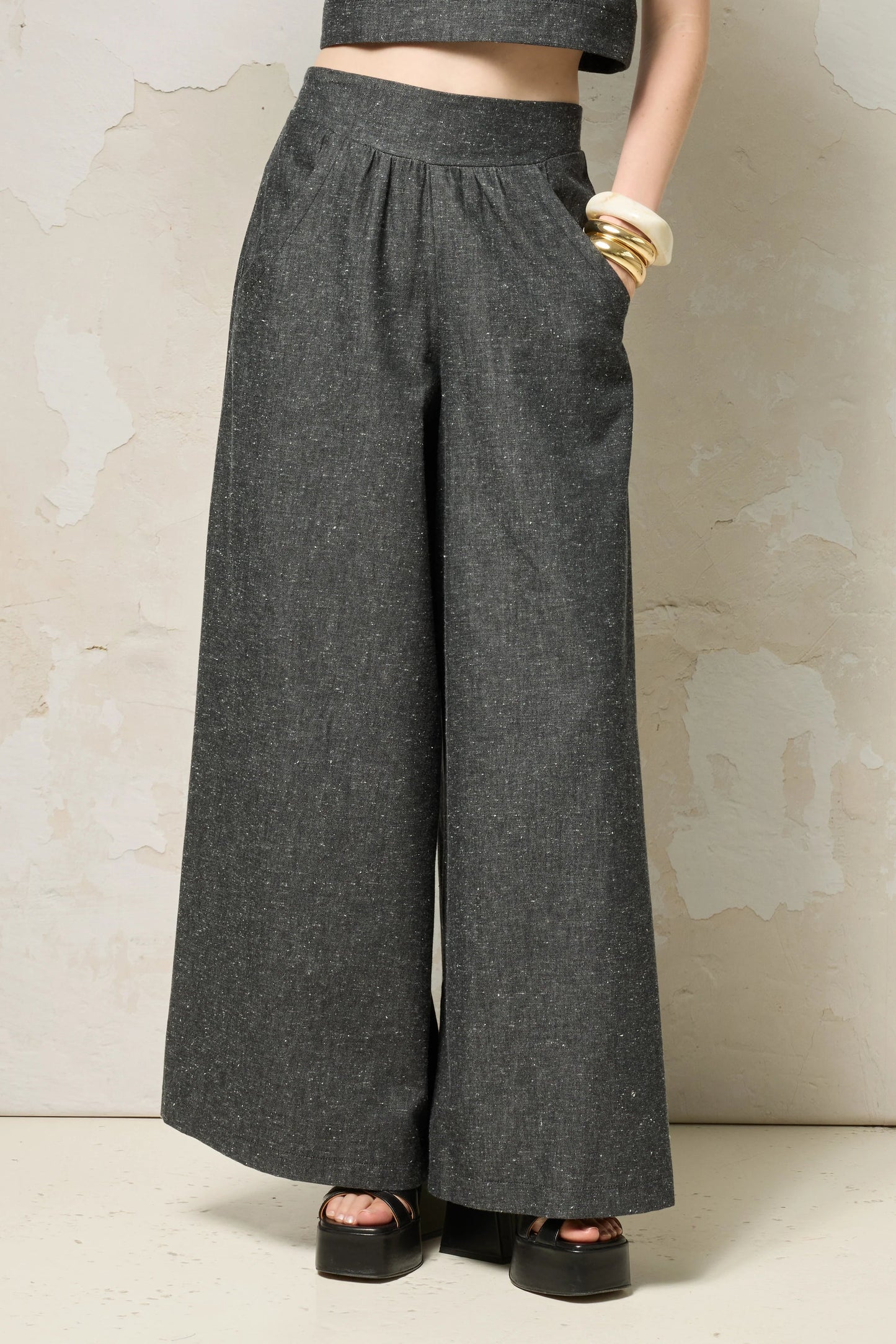 Waist down view of a woman wearing the Rama Pants by Cokluch in Black Speckle, textured denim pants with wide legs and a smooth elastic waistband. She is wearing them with a matching top and standing in front of a grey wall. 