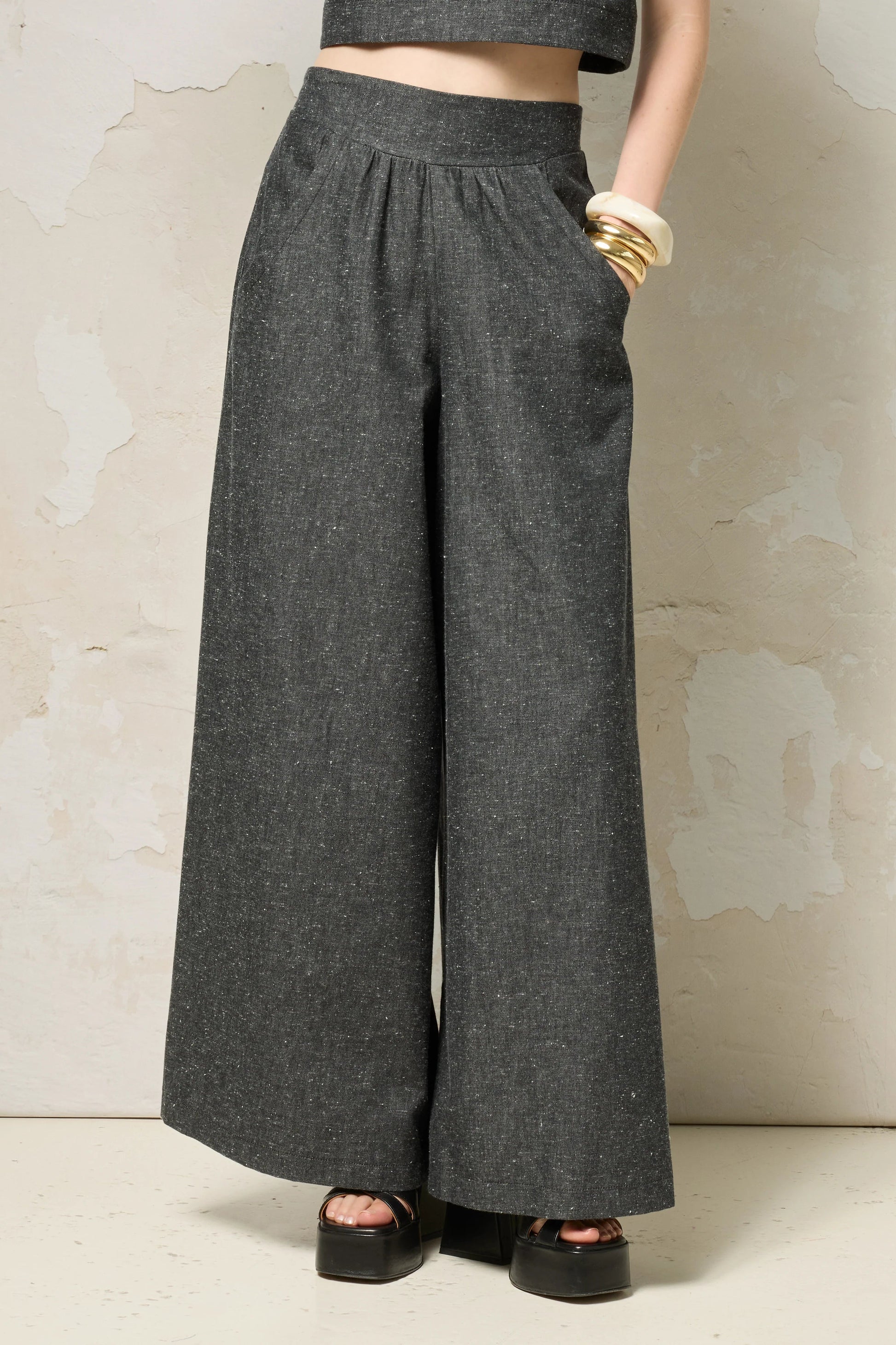 Waist down view of a woman wearing the Rama Pants by Cokluch in Black Speckle, textured denim pants with wide legs and a smooth elastic waistband. She is wearing them with a matching top and standing in front of a grey wall. 