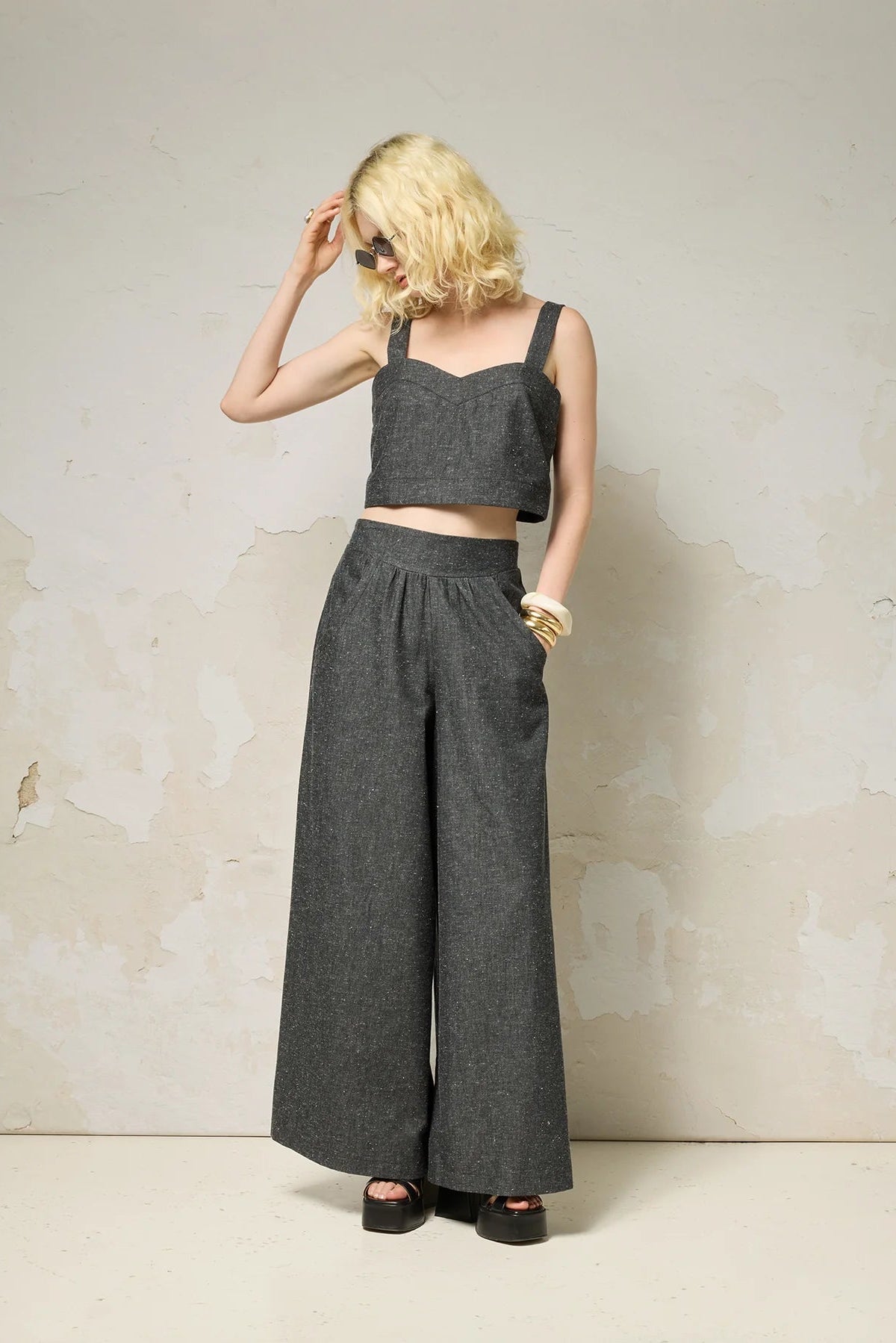 A woman wearing the Rama Pants by Cokluch in Black Speckle, textured denim pants with wide legs and a smooth elastic waistband. She is wearing them with a matching top and standing in front of a grey wall. 
