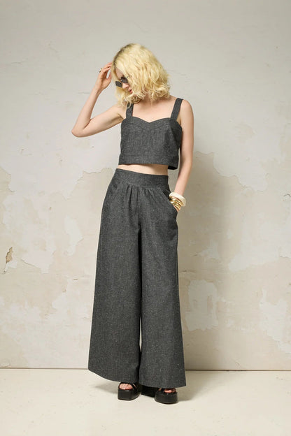 A woman wearing the Rama Pants by Cokluch in Black Speckle, textured denim pants with wide legs and a smooth elastic waistband. She is wearing them with a matching top and standing in front of a grey wall. 