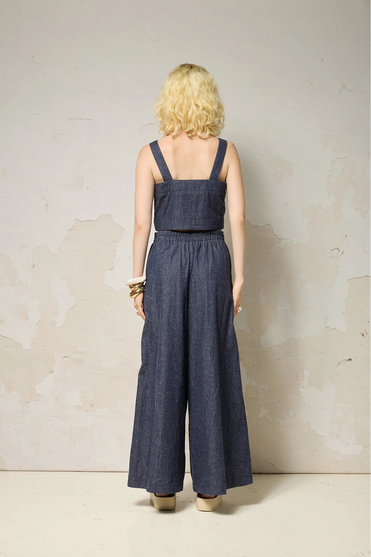 Back view of a woman wearing the Rama Pants by Cokluch in Indigo Speckle, textured denim pants with wide legs and a smooth elastic waistband. She is wearing them with a matching top and standing in front of a grey wall. 