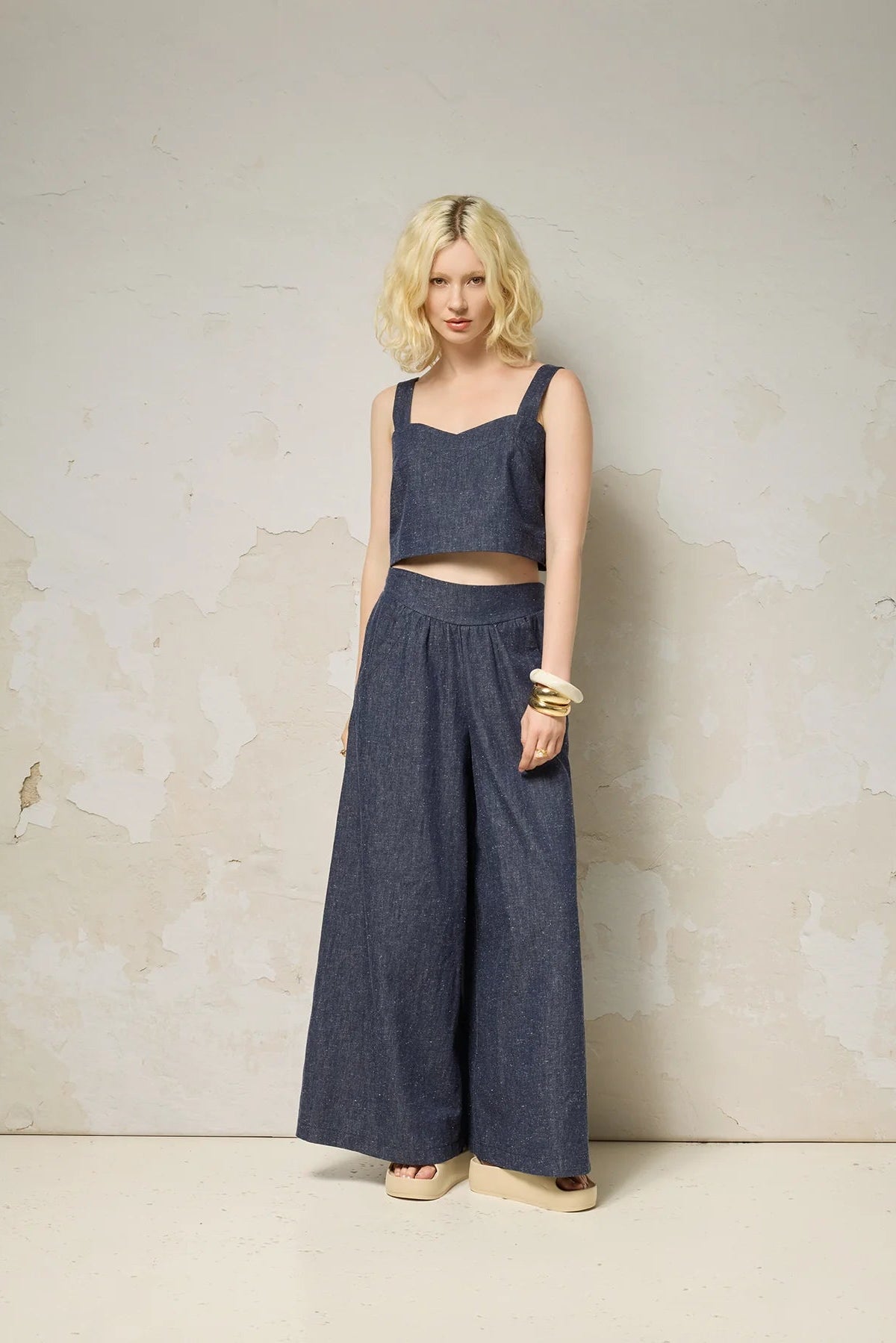 A woman wearing the Rama Pants by Cokluch in Indigo Speckle, textured denim pants with wide legs and a smooth elastic waistband. She is wearing them with a matching top and standing in front of a grey wall. 