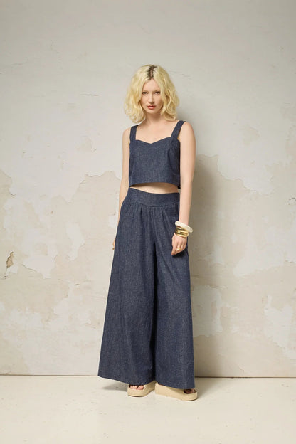  A woman wearing the Rama Pants by Cokluch in Indigo Speckle, textured denim pants with wide legs and a smooth elastic waistband. She is wearing them with a matching top and standing in front of a grey wall. 